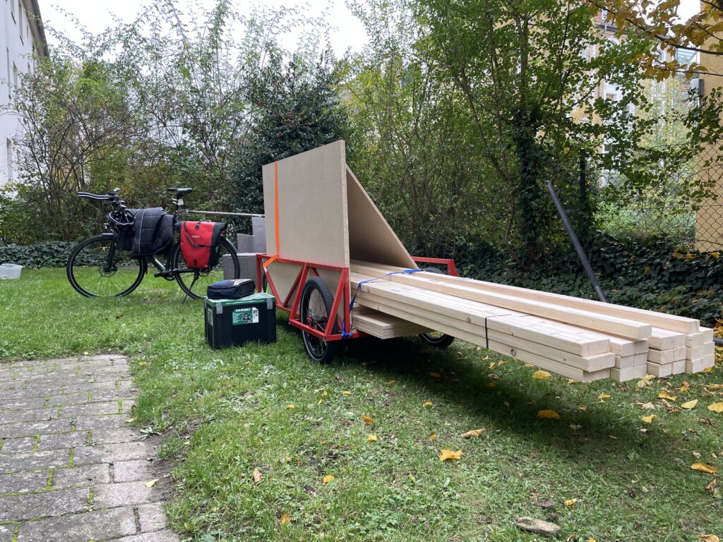 Zu sehen ist ein großer Fahrradanhänger, der an einem Fahrrad hängt. Auf diesem Anhänger sind ein paar Holzlatten mit einer Länge von ca. 3 m und ein paar Holzplatten mit einer Grundfläche von über 1 m² festgespannt.