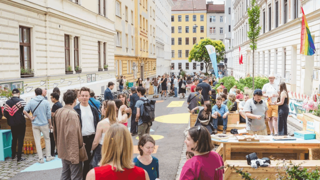 Ein Bild von der Galileistraße in Wien. Viele Menschen befinden sich auf der Straße und unterhalten sich. Es wurden in den Seitenräumen Holzstrukturen aufgebaut, die von dem Menschen als Tische und Bänke genutzt werden können.