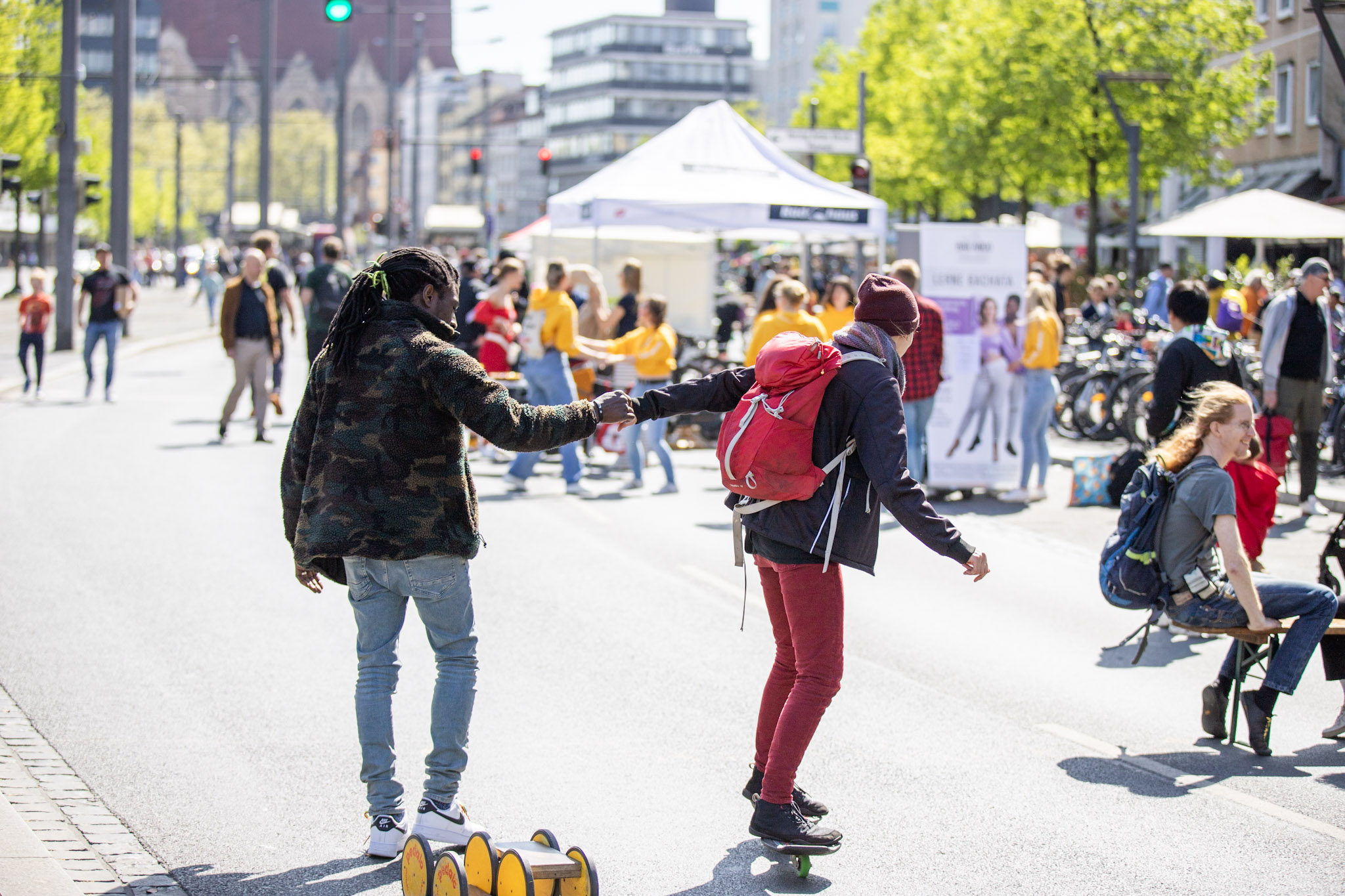 Im Mittelpuntk des Bildes sind zwei Menschen zu sehen. Die eine Person hilft der anderen Person beim Waveboard fahren. Dahinter sind weitere Menschen zu sehen, die sitzen und sich unterhalten und wiederum andere Menschen, die tanzen. Die Sonne scheint und die Menschen sehen glücklich aus.