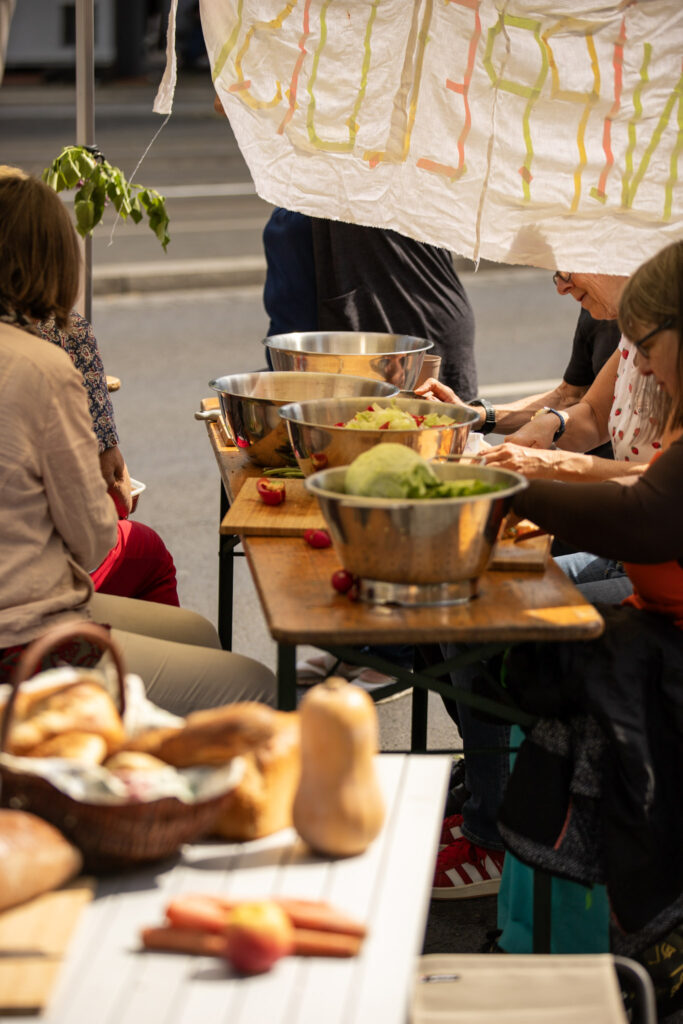 Es wird auf Bierzeltgarnituren gekocht. gerade schnippeln Menschen am Gemüse. Im Hintergrund hängt ein Banner, auf dem der Begriff "Schnippelzeit" gelesen werden kann. Das Kochen findet auf dem Bohlweg statt.