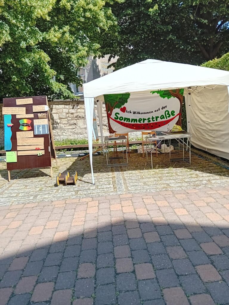 Zu sehen ist ein Pavillon, in dem ein Banner hängt, auf dem "Herzlich Willkommen auf der Sommerstraße" steht. Es ist das gleiche Banner, wie auch schon bei der Sommerstraße 2025 auf dem Bohlweg hing. In dem Pavillon sind Tische und Stühle zu sehen. Links davon steht ein kleines Pedalo auf dem Boden, daneben steht ein menschengroßer Holzaufsteller, auf dem das "Sommerstraßen Quiz" abgebildet ist. Darauf sind verschiedene Fragen spielerisch aufgebaut, wie hoch beipsielsweise das Wasser auf dem Bohlweg beim Starkregen von 2023 stand der wieviele Fahrräder auf einen Pkw-Stellplatz passen.