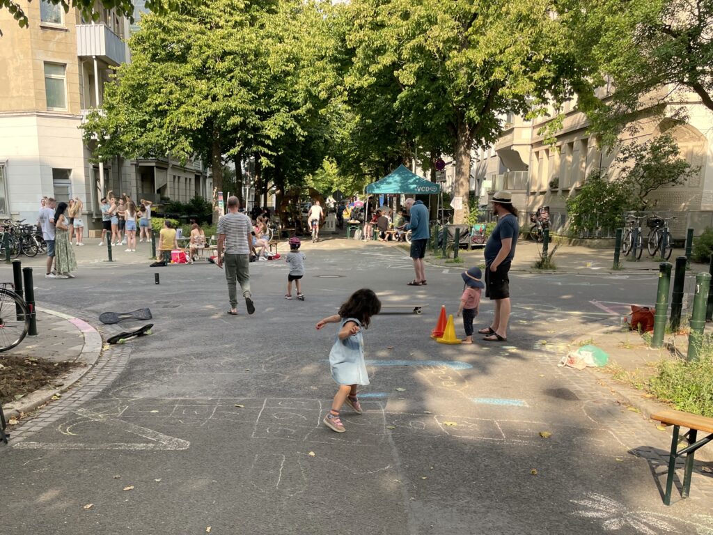 Eine Kreuzung einer Wohnstraße. Es befinden sich mehrere Menschen auf der Straße. Dabei fallen vor allem spielende Kinder auf. Weiter entfernt ist eine Tanzgruppe zu erkennen.