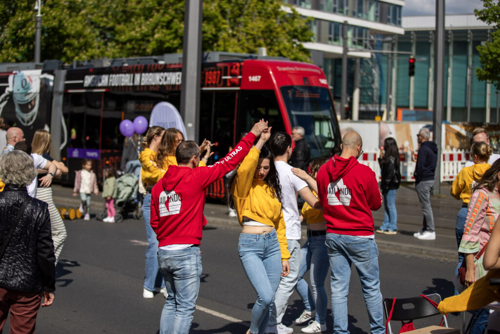 Menschen Tanzen auf dem Bohlweg. Weiter weg sieht man spielende Kinder und eine Straßenbahn, die an dem Geschehen vorbeifährt.
