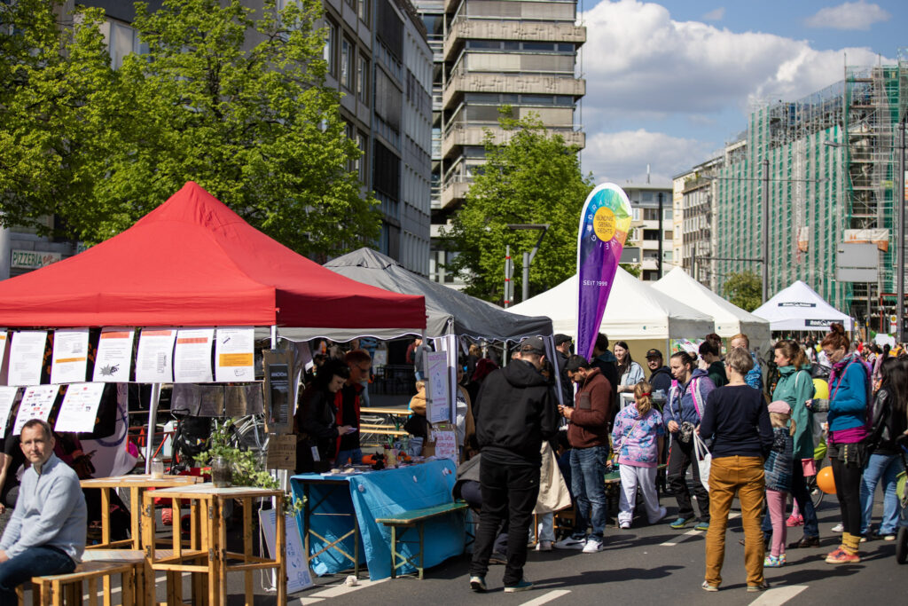 Man sieht viele Infostände (Pavillons) auf der Straße und Menschen, die sich unterhalten.